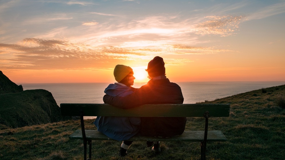 Two people watch the sunset from a bench on Howth Head near Dublin, overlooking the Irish Sea on a romantic evening.