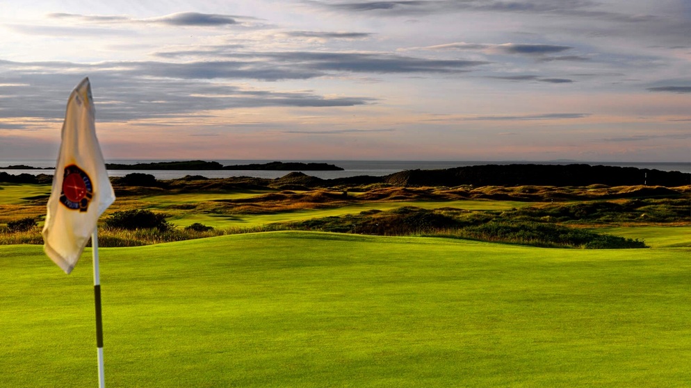 Green fairways stretch towards the coast at dusk, with a golf flag in the foreground and calm waters under a pastel sky.