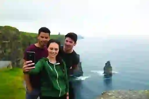 Three friends take a selfie at the edge of the Cliffs of Moher, with sheer sea cliffs rising from the Atlantic below.