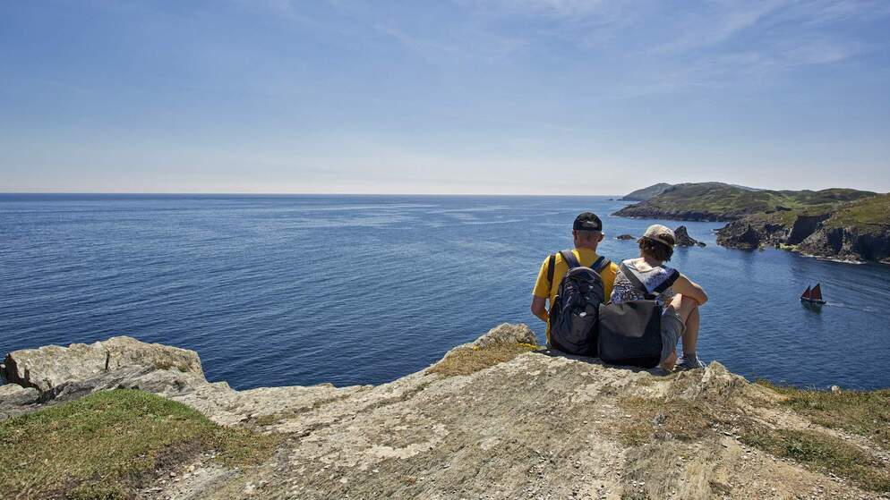 Two hikers sit on cliffs on the coast of County Kerry, overlooking the Atlantic Ocean as a sailboat passes below.