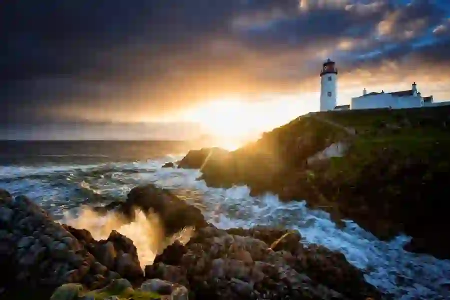 Fanad Head Lighthouse on a rocky headland with waves breaking below at sunset on the Atlantic.
