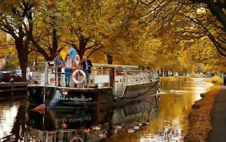 Group of people on a canal boat in Dublin, surrounded by autumn trees and ducks swimming nearby.