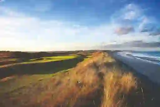 Dramatic coastal view of golden dunes and green fairways beside a sweeping sandy beach under a bright blue sky.