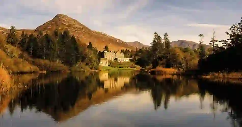 Het historische Ballynahinch Castle Hotel in county Galway weerspiegelt in een rustig meer, met een berg op de achtergrond, onder een gouden lucht.