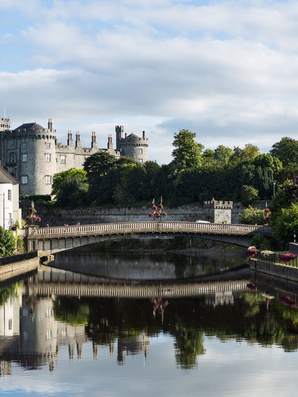 Stone bridge over the River Nore facing Kilkenny Castle and old town buildings surrounded by trees and flowers.
