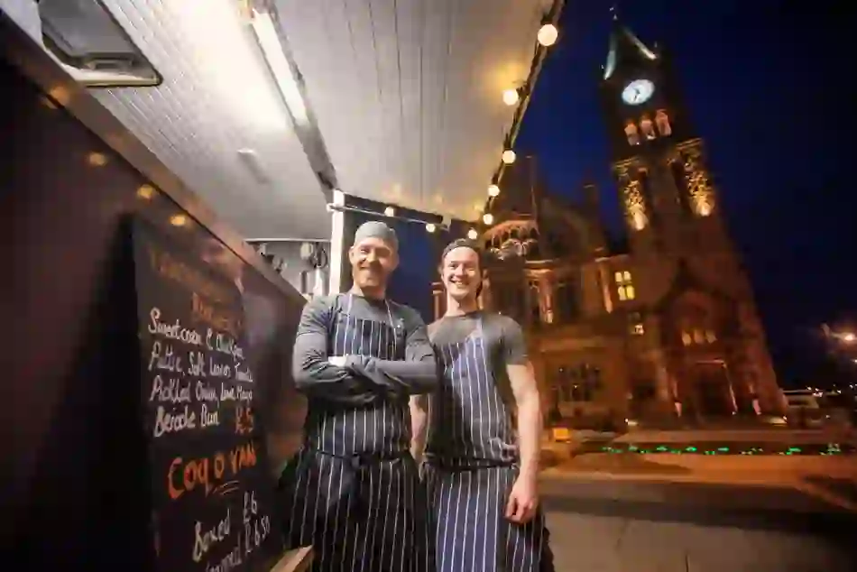Two smiling street food vendors in striped aprons at a night market in Derry~Londonderry, with Guildhall clock tower behind.