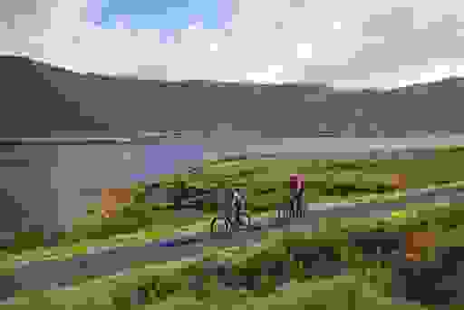 Two cyclists ride a lakeside path in Mayo, golden bogland and mountain slopes reflecting in still waters.