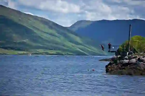 Two people jump off a wooden platform into Killary Fjord, with steep green mountains rising behind in County Galway.