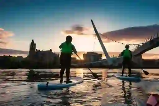 Two paddleboarders at sunset on the River Foyle pass under Derry~Londonderry’s Peace Bridge with Guildhall in view.