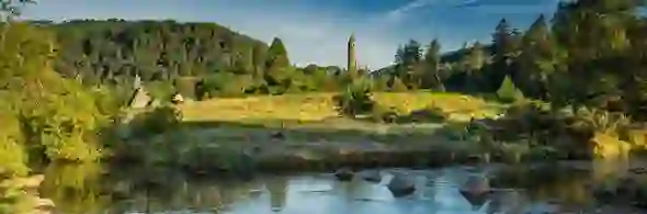 Glendalough Lake Walk in County Wicklow with monastic round tower rising above trees beside a calm river.
