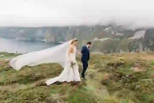 Bride and groom walking along Slieve League Cliffs in Donegal, with ocean and mist-covered cliffs in the background.