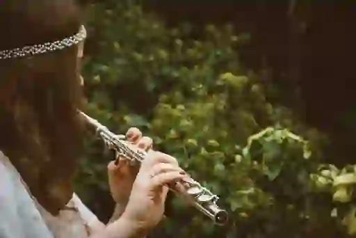 Woman wearing pearl headband playing silver flute outdoors, surrounded by green foliage.