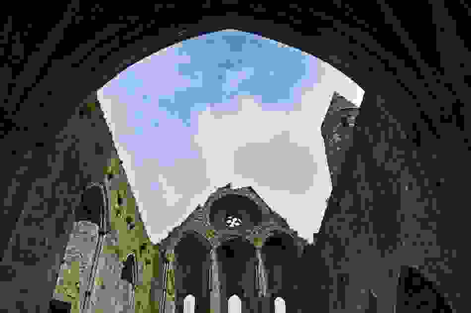 Interior ruins of the Rock of Cashel, County Tipperary, framed by Gothic arches and a round tower against blue sky.