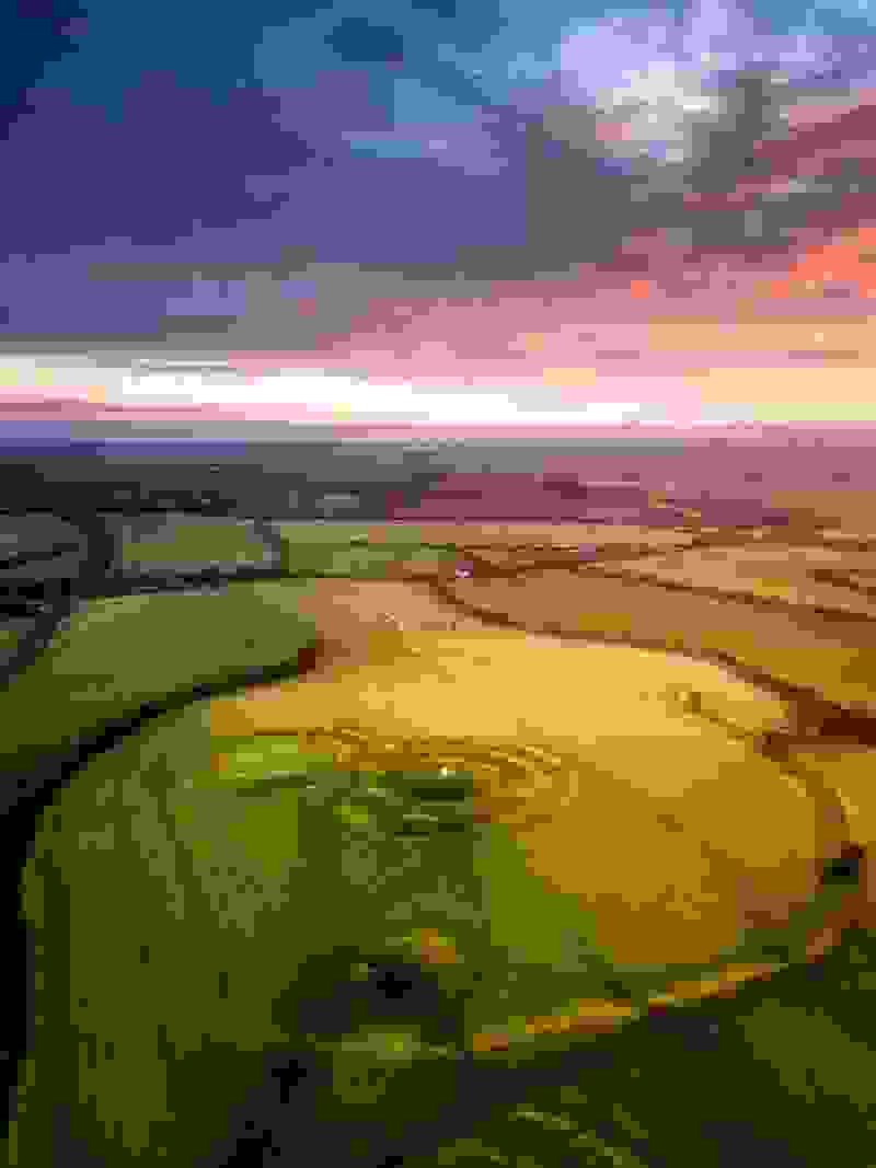 Aerial view of the Hill of Tara at sunrise, with patchwork fields glowing under a pink and orange sky.