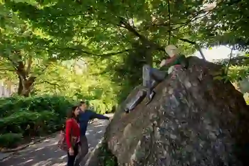 Couple gazes at Oscar Wilde statue in Merrion Square, surrounded by trees and rich greenery.