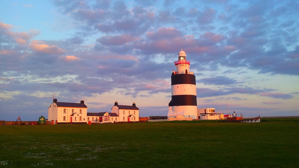 Hook Lighthouse with bold black-and-white bands at sunset, beside white cottages under a purple-pink coastal sky.