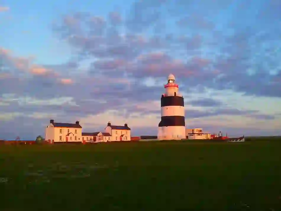 Hook Lighthouse with bold black-and-white bands at sunset, beside white cottages under a purple-pink coastal sky.