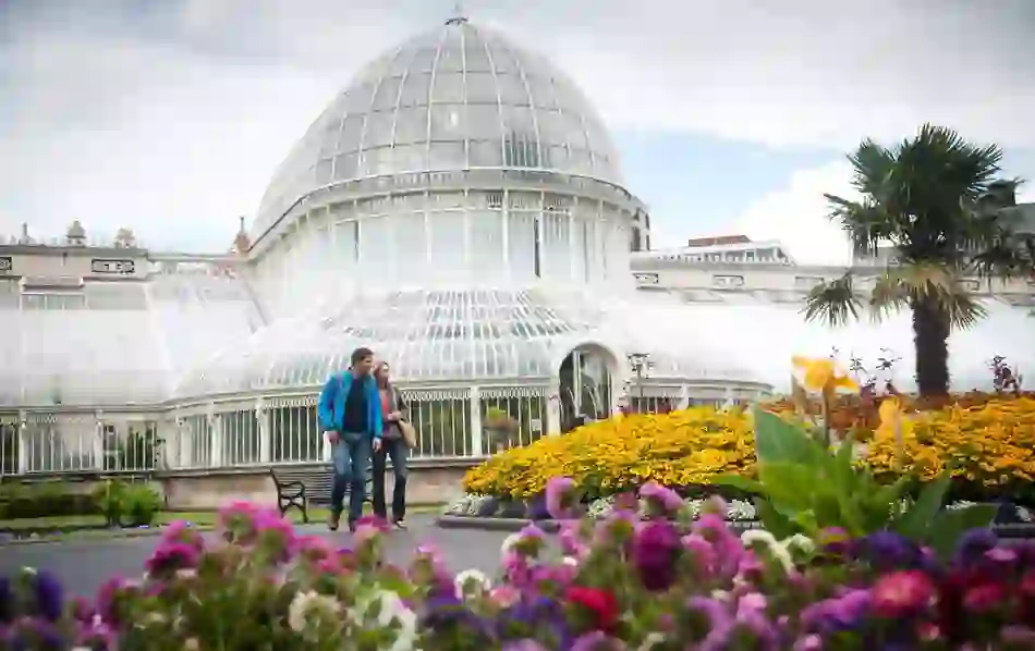 Couple walks past flowerbeds and palm trees in front of the domed glass Palm House in Belfast Botanic Gardens.