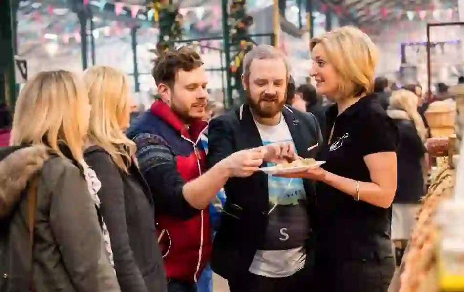 People sample food from a smiling vendor at a busy stall in St George’s Market, decorated with bunting.