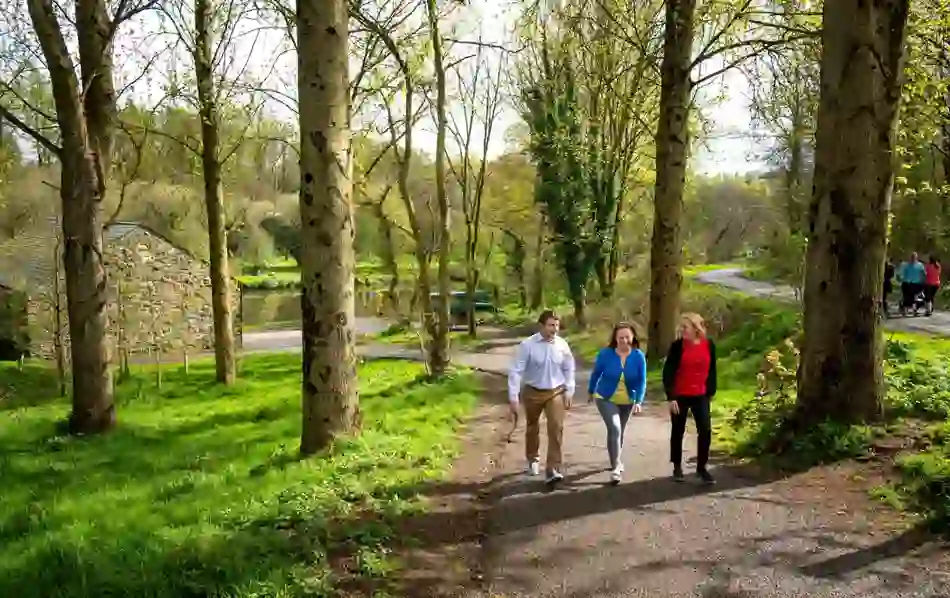People walking along the River Lagan towpath through trees and past a stone building.
