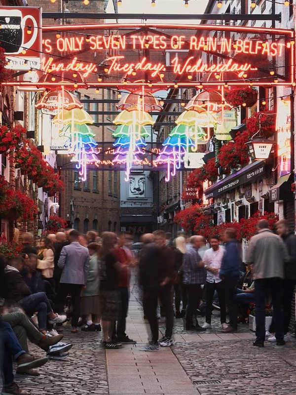 Neon umbrellas light up a lively street in Belfast’s Cathedral Quarter, decorated with flowers.
