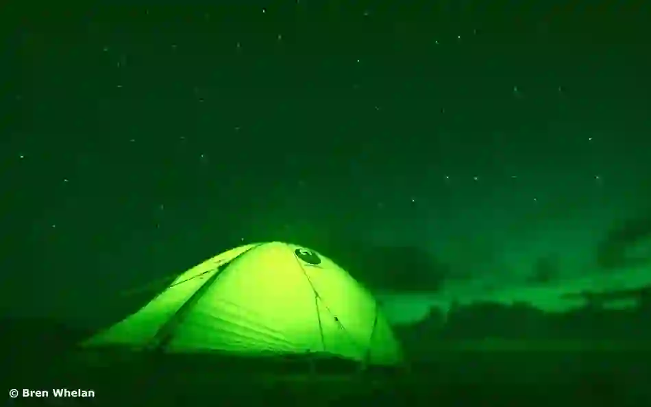 Green tent glows beneath a starry night sky as the Aurora Borealis shimmers above Malin Head’s coastal landscape.