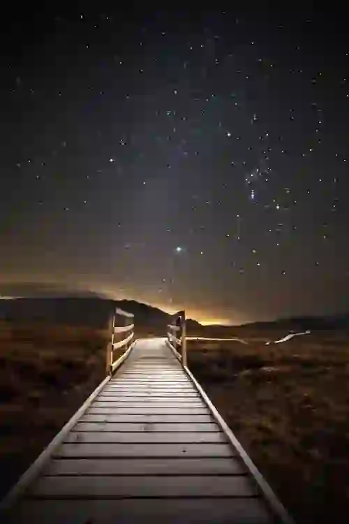 Wooden boardwalk leads into Ballycroy National Park under a star-filled night sky with Orion visible above distant hills.
