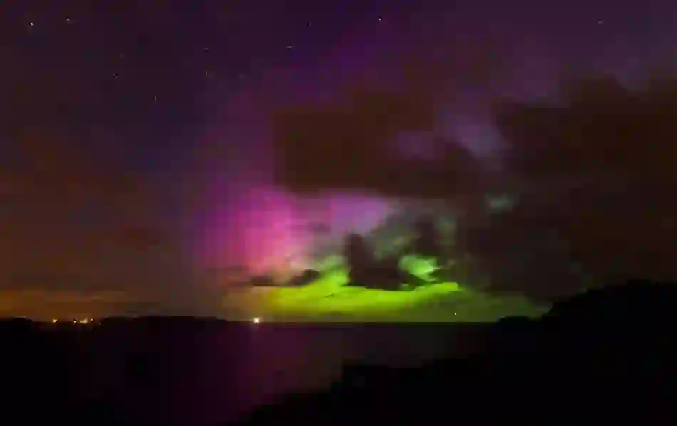 Northern Lights in pink and green hues illuminate clouds over the sea near Fort Dunree’s rugged coastline at night.