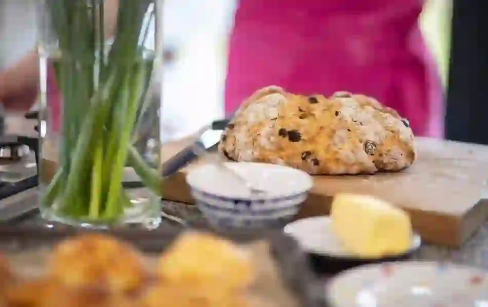 Freshly baked Irish soda bread with raisins on a wooden board, served with butter and bowls on a kitchen table.