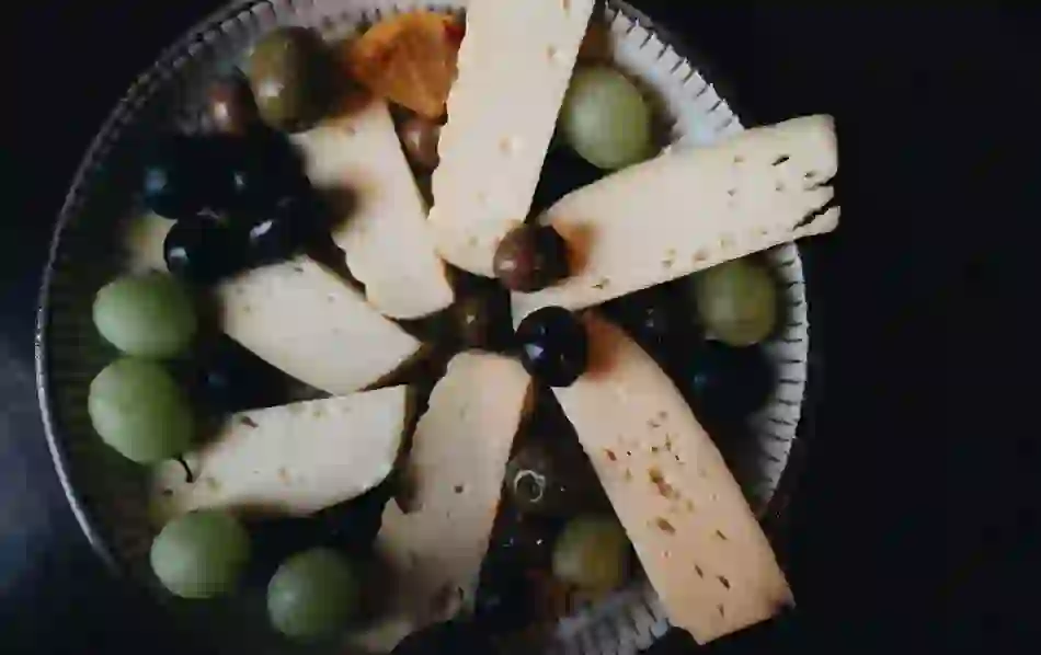 Cheese platter with sliced semi-hard cheese, green and black grapes, olives and orange on a decorative plate.