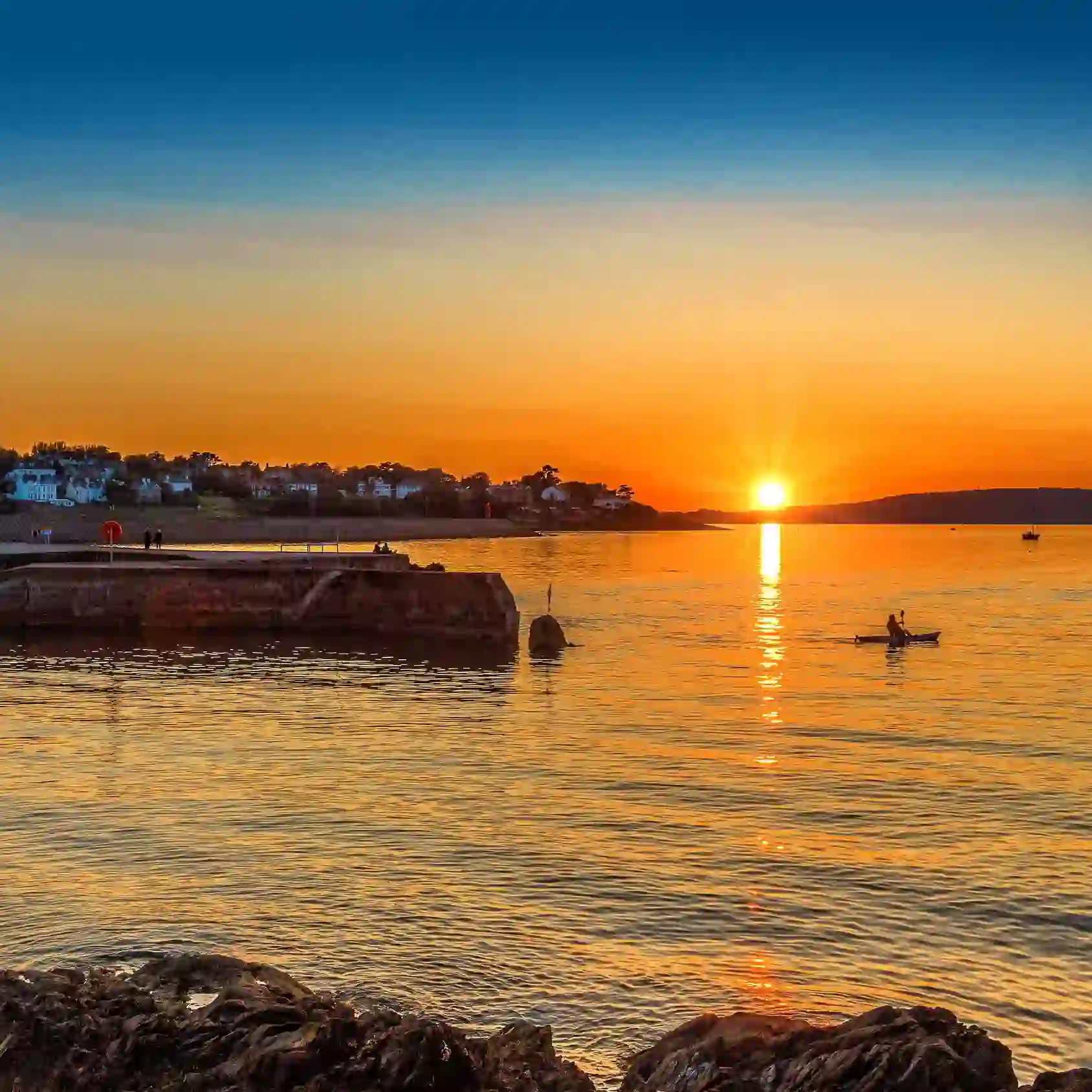 Sunset over Bangor marina with glowing orange sky, calm water, pier and a lone kayaker on the bay.