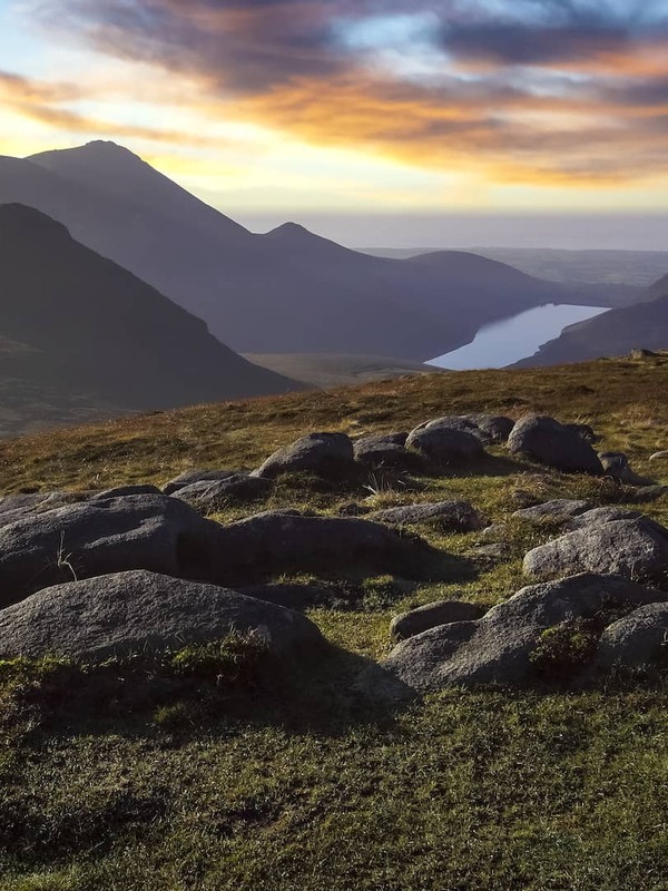 Sunrise over the Mourne Mountains in County Down with rocky peaks, a lakeside valley and a tent overlooking the landscape.