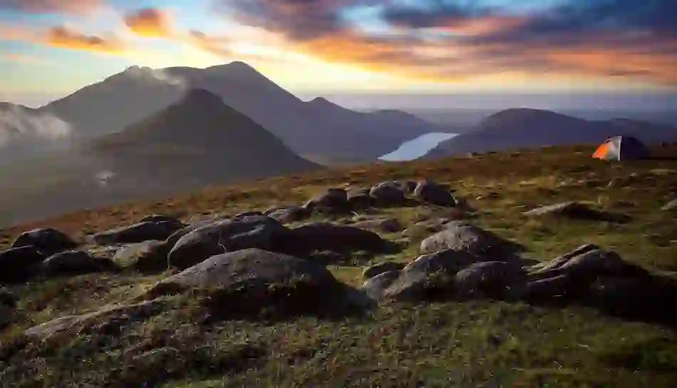 Sunrise over the Mourne Mountains in County Down with rocky peaks, a lakeside valley and a tent overlooking the landscape.