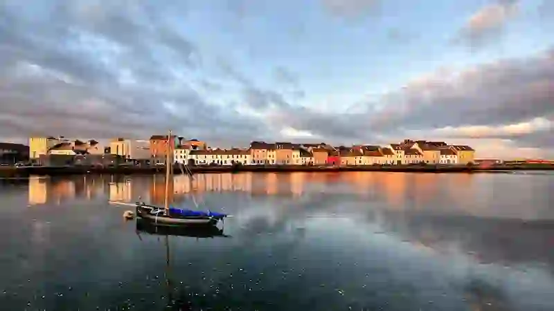 Galway city's Long Walk and The Claddagh at sunset with colourful houses reflected in calm water.