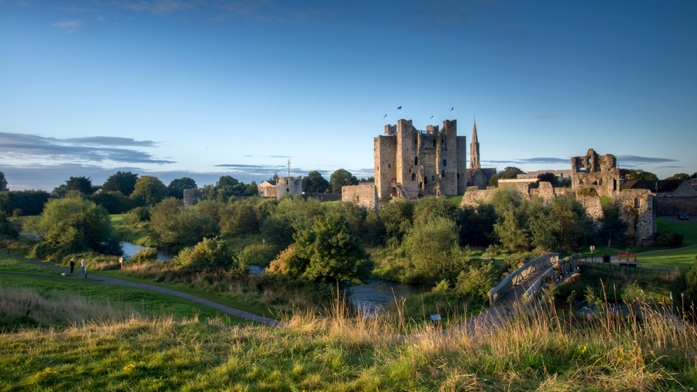 Trim Castle rises above the River Boyne, with a footbridge, green parkland and blue evening sky in County Meath.