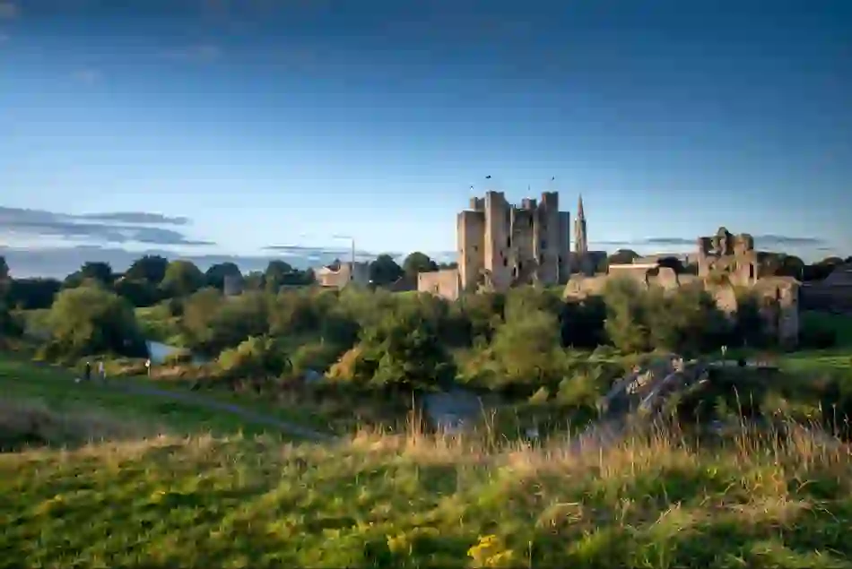 Trim Castle rises above the River Boyne, with a footbridge, green parkland and blue evening sky in County Meath.