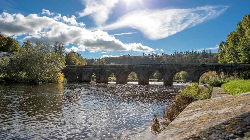 Stone arch bridge over the River Nore in Inistioge, County Kilkenny, with trees and blue sky reflected in the water.