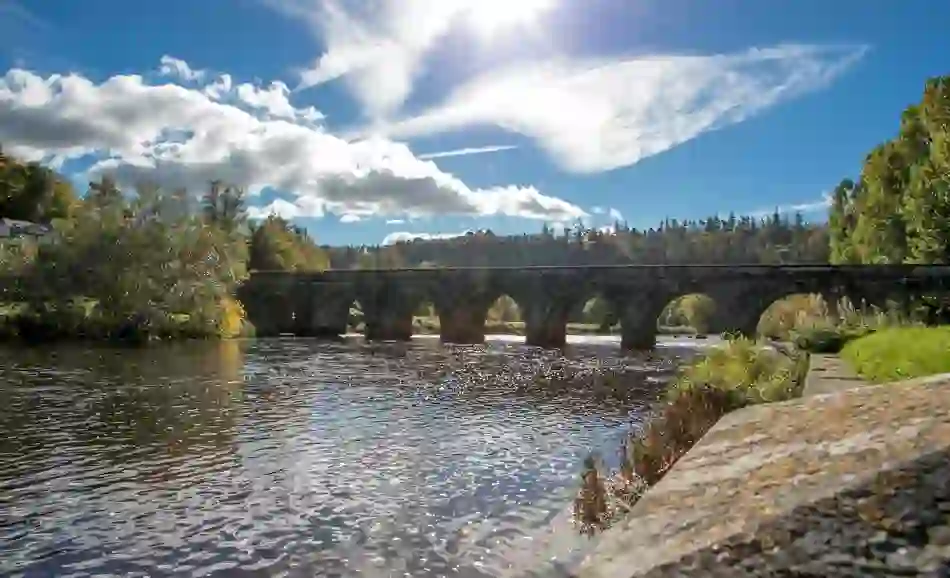 Stone arch bridge over the River Nore in Inistioge, County Kilkenny, with trees and blue sky reflected in the water.