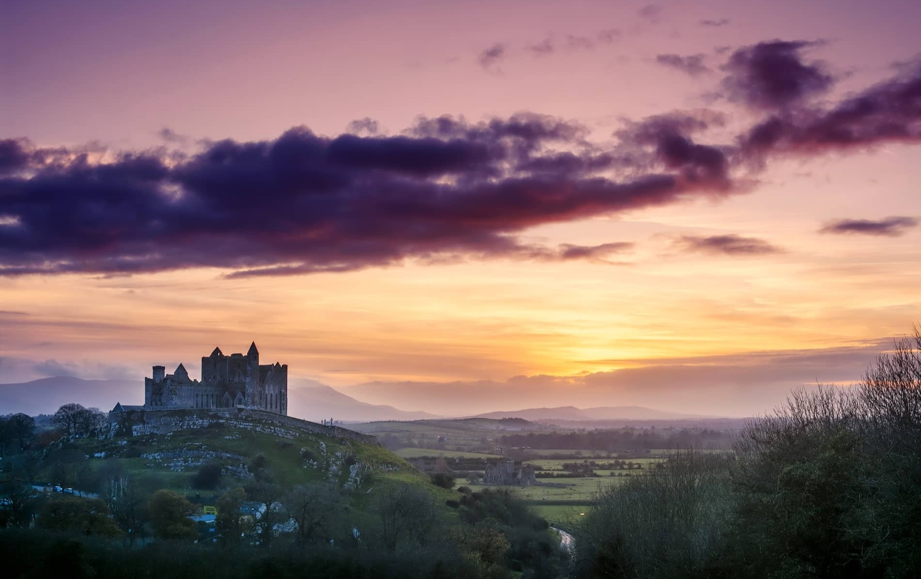Rock of Cashel silhouetted at sunset over rolling countryside in County Tipperary, with vibrant pink and orange skies.
