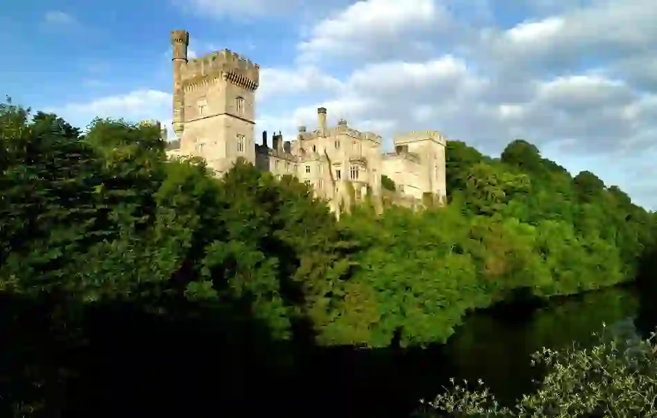 Lismore Castle, County Waterford, overlooking the River Blackwater, surrounded by lush green woodland under blue sky.