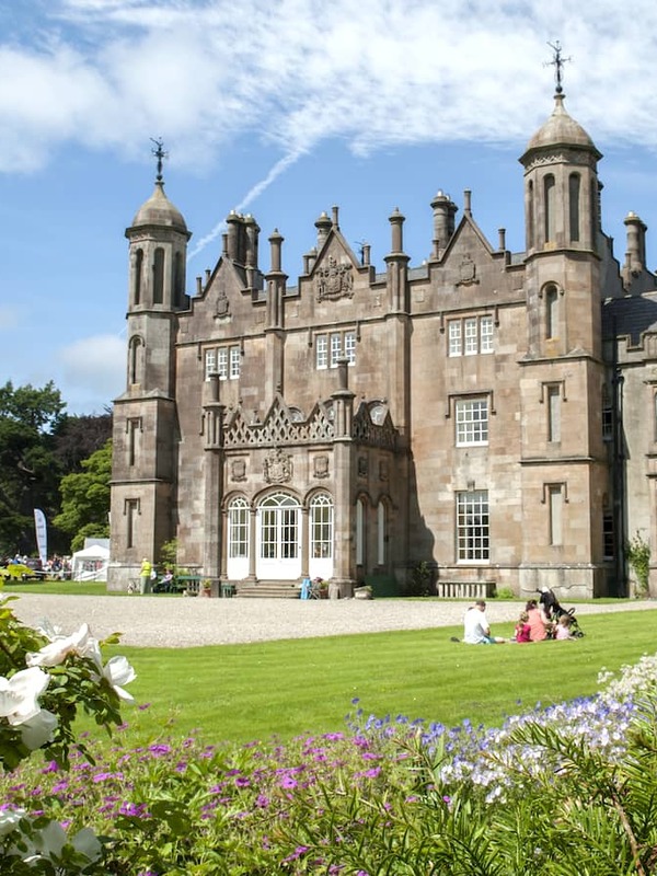 Glenarm Castle and gardens in County Antrim, with visitors relaxing on the lawn beside colourful flowerbeds.