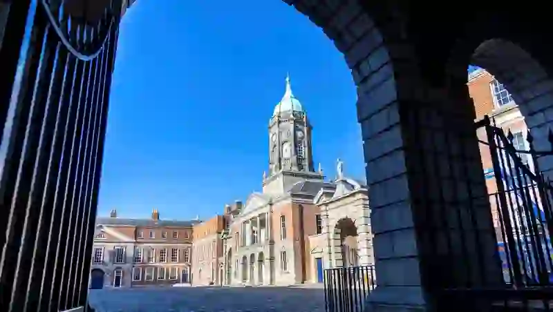 Clock tower and courtyard of historic Dublin Castle viewed through iron gates on a clear day with blue skies.