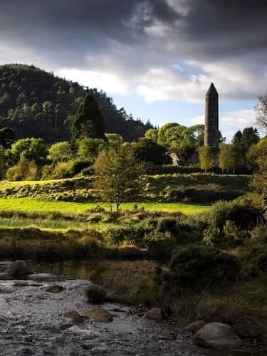 Sunlight breaks through clouds over Glendalough's monastic ruins and round tower, nestled in a lush green valley by a flowing stream.