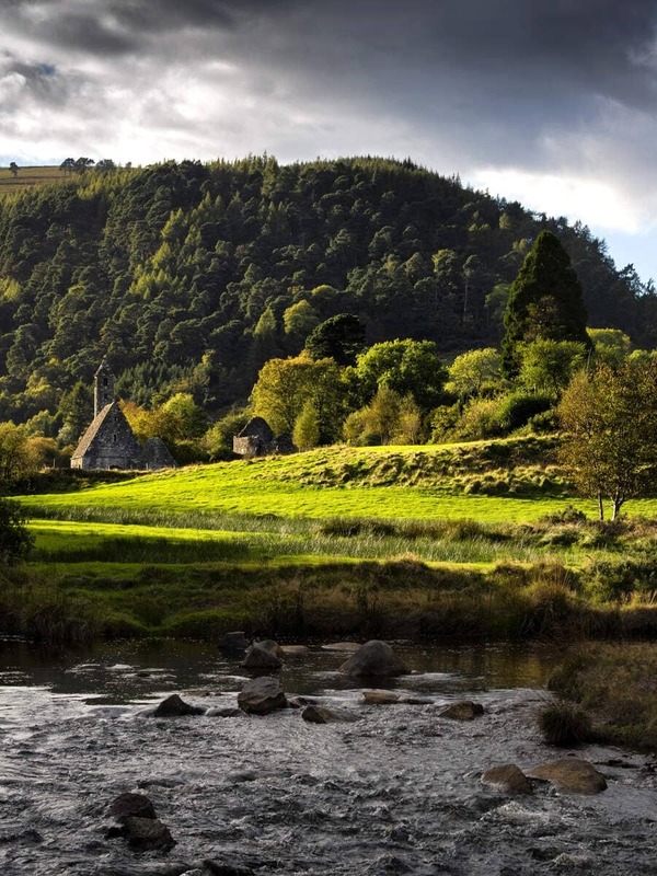 Sunlight breaks through clouds over Glendalough's monastic ruins and round tower, nestled in a lush green valley by a flowing stream.