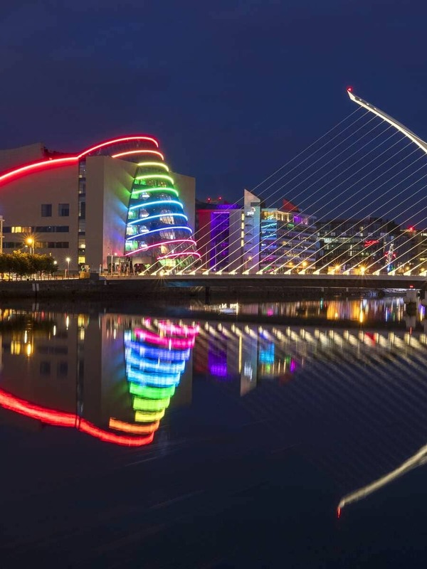 Colourfully lit Convention Centre and Samuel Beckett Bridge reflected in the River Liffey at night.
