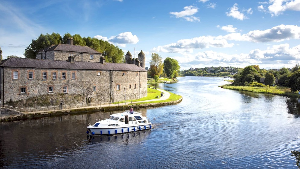 A white cruiser boat passes Enniskillen Castle on the River Erne under a bright blue sky in County Fermanagh, Northern Ireland.