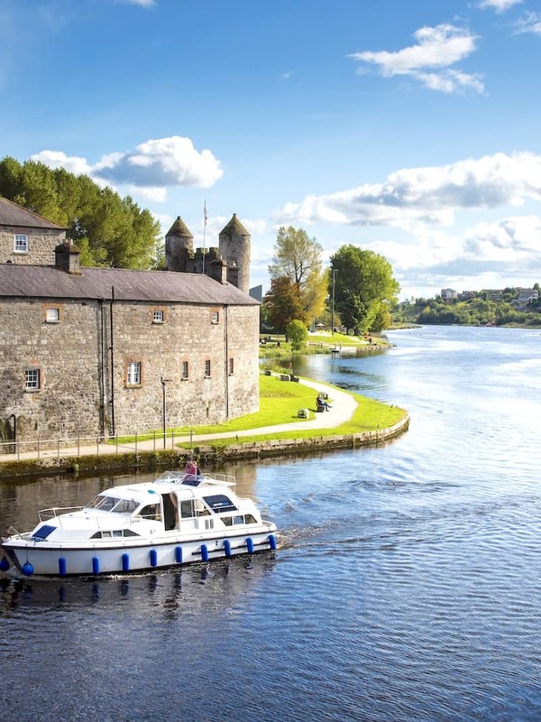 A white cruiser boat passes Enniskillen Castle on the River Erne under a bright blue sky in County Fermanagh, Northern Ireland.