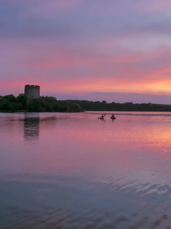 Two kayakers paddle across Lough Oughter in County Cavan at sunset, with a castle tower silhouetted against the pink sky.