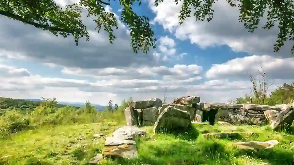 Prehistoric stone tomb surrounded by green grass and trees under a partly cloudy sky at Cavan Burren Park, County Cavan.