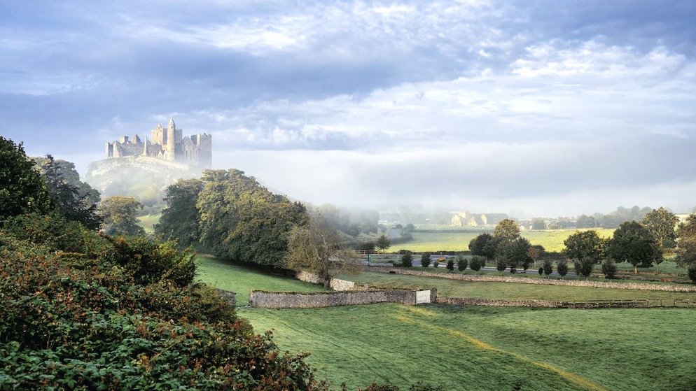 Rock of Cashel rising above green farmland of County Tipperary, with medieval stone buildings framed by misty morning light.